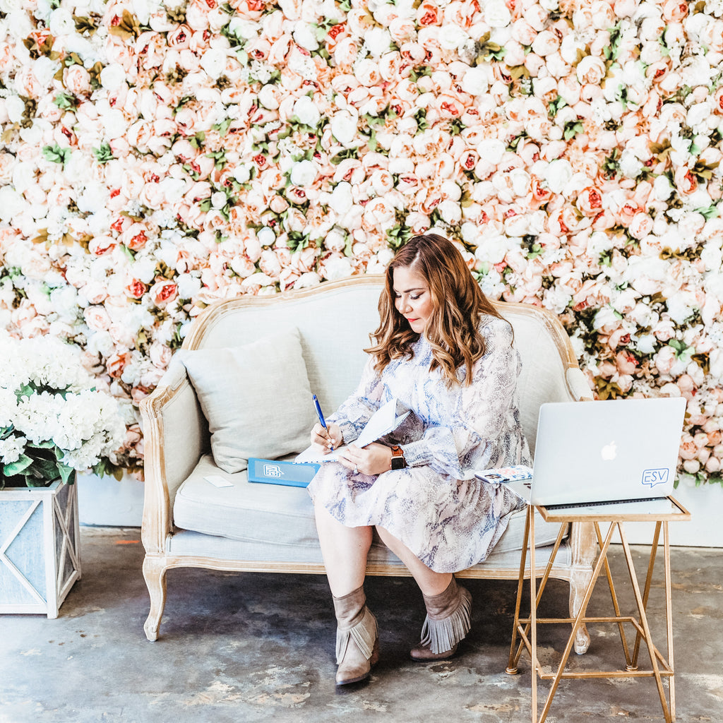 A woman sits on a cream sofa, writing in a notebook about her ESV Creative 90-Minute Social Media Intensive strategy, with a laptop beside her. She is in front of a wall covered in pink and white flowers.