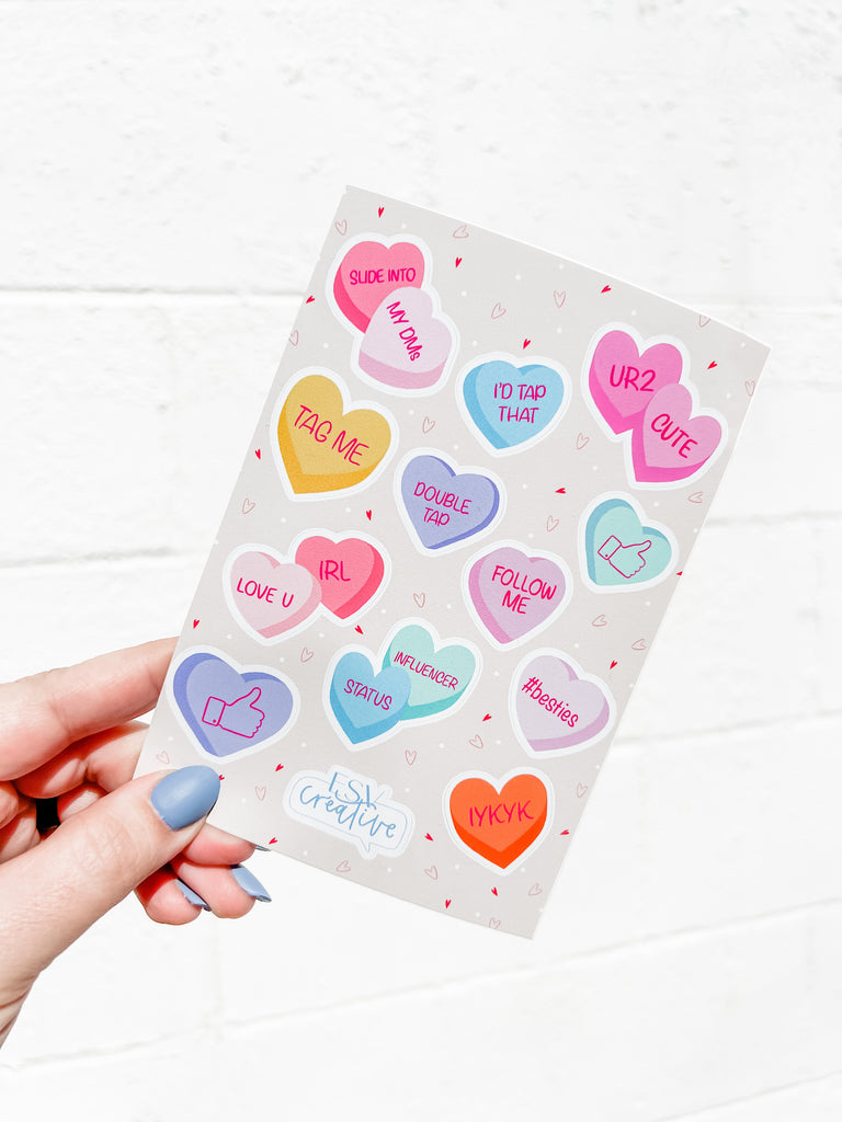 A hand holding a card against a white wall, featuring various colorful waterproof vinyl stickers shaped like hearts with playful social media-themed phrases like "follow me," "tag me," and emojis from the ESV Creative Social Media Conversation Heart Sticker Sheet.