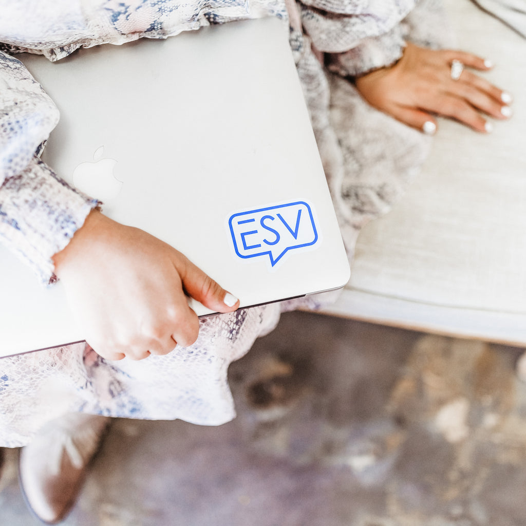 A person sitting on a floor with a closed laptop in their lap, showing a sticker promoting their ESV Creative social media presence. They are wearing a floral dress and brown boots.